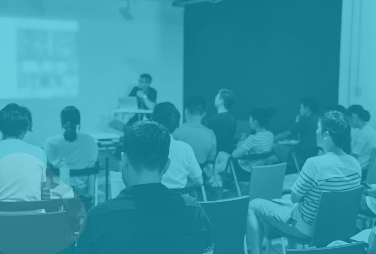 A group of students sits in a classroom listening to a teacher who is presenting at the front of the room. The entire image is overlaid with a blue tint.